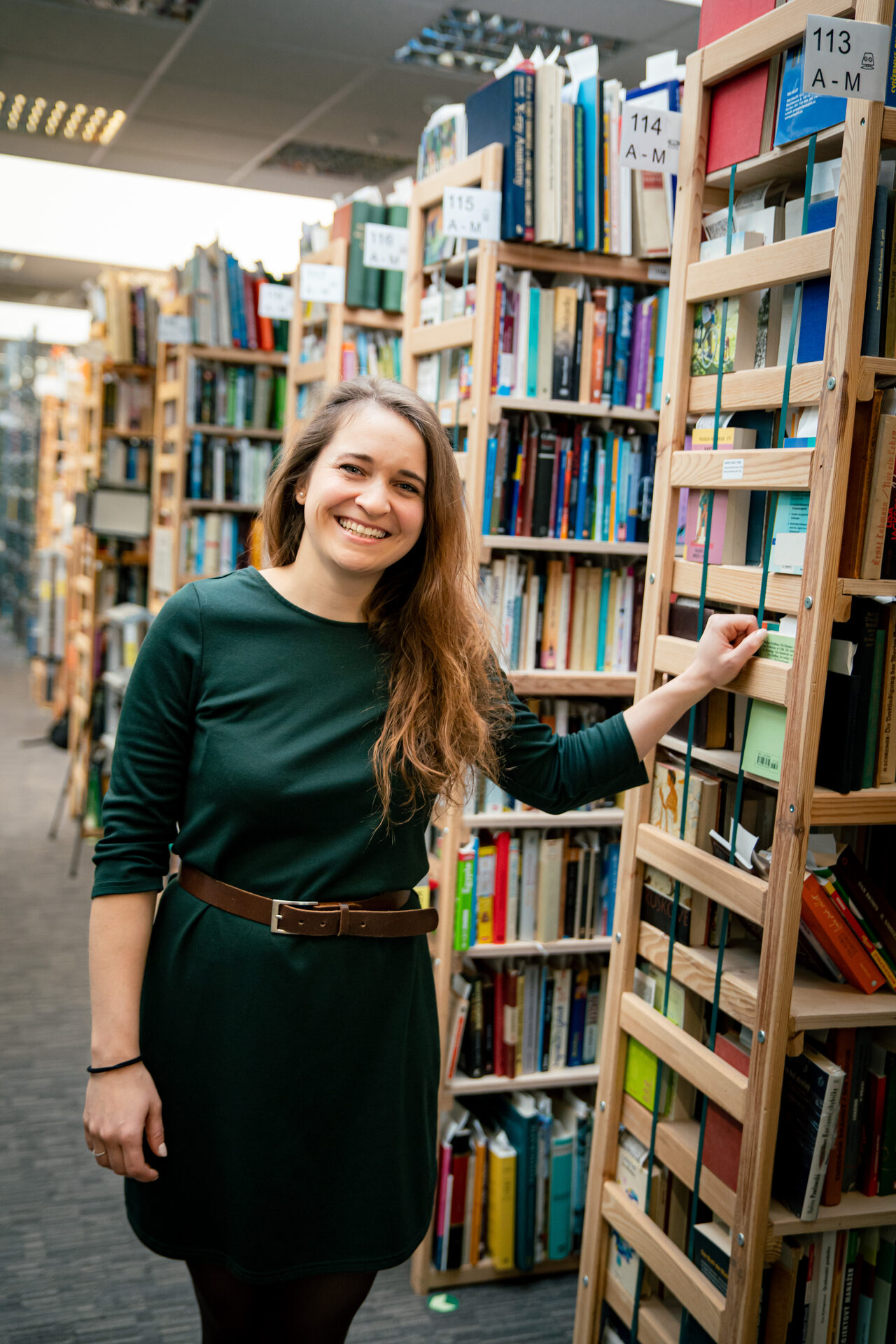 Kateřina Nushartová, Head of HR ve společnosti Knihobot, stojí s úsměvem u vysokého dřevěného regálu plného knih. Na sobě má tmavě zelené šaty s hnědým páskem. Fotografie vznikla během natáčení rozhovoru pro sérii HR Vibecheck od agentury VERYGUD.