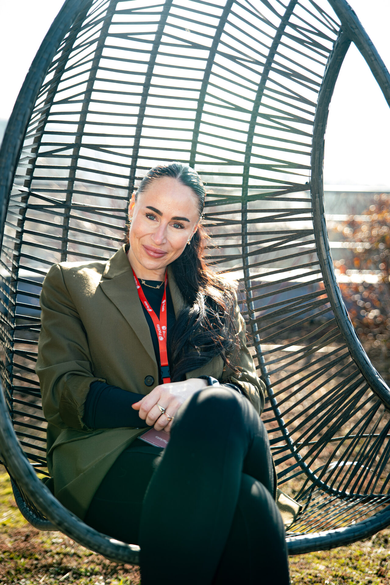 Caroline S. Karda, Group Head of Talent Acquisition ve společnosti Packeta, sedí venku v černém proutěném závěsném křesle a usmívá se do objektivu. Na sobě má tmavě zelené sako a na krku červenou šňůrku Packeta. Foceno pro sérii HR Vibecheck od agentury VERYGUD.