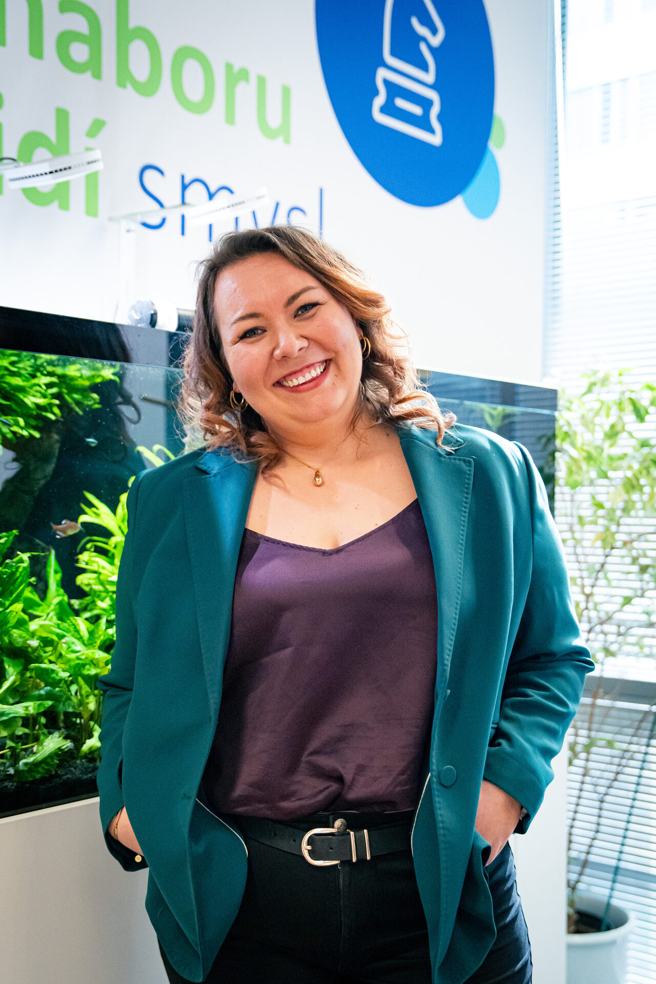 Tereza Kundert Silbernáglová, Head of HR Partners ve společnosti mBlue, se usmívá do kamery s rukama v kapsách. Na sobě má zelené sako a vínovou halenku. Stojí v prosvětlené kanceláři vedle velkého akvária s rostlinami, na zdi v pozadí je vidět firemní grafika. Foceno pro sérii HR Vibecheck od agentury VERYGUD.