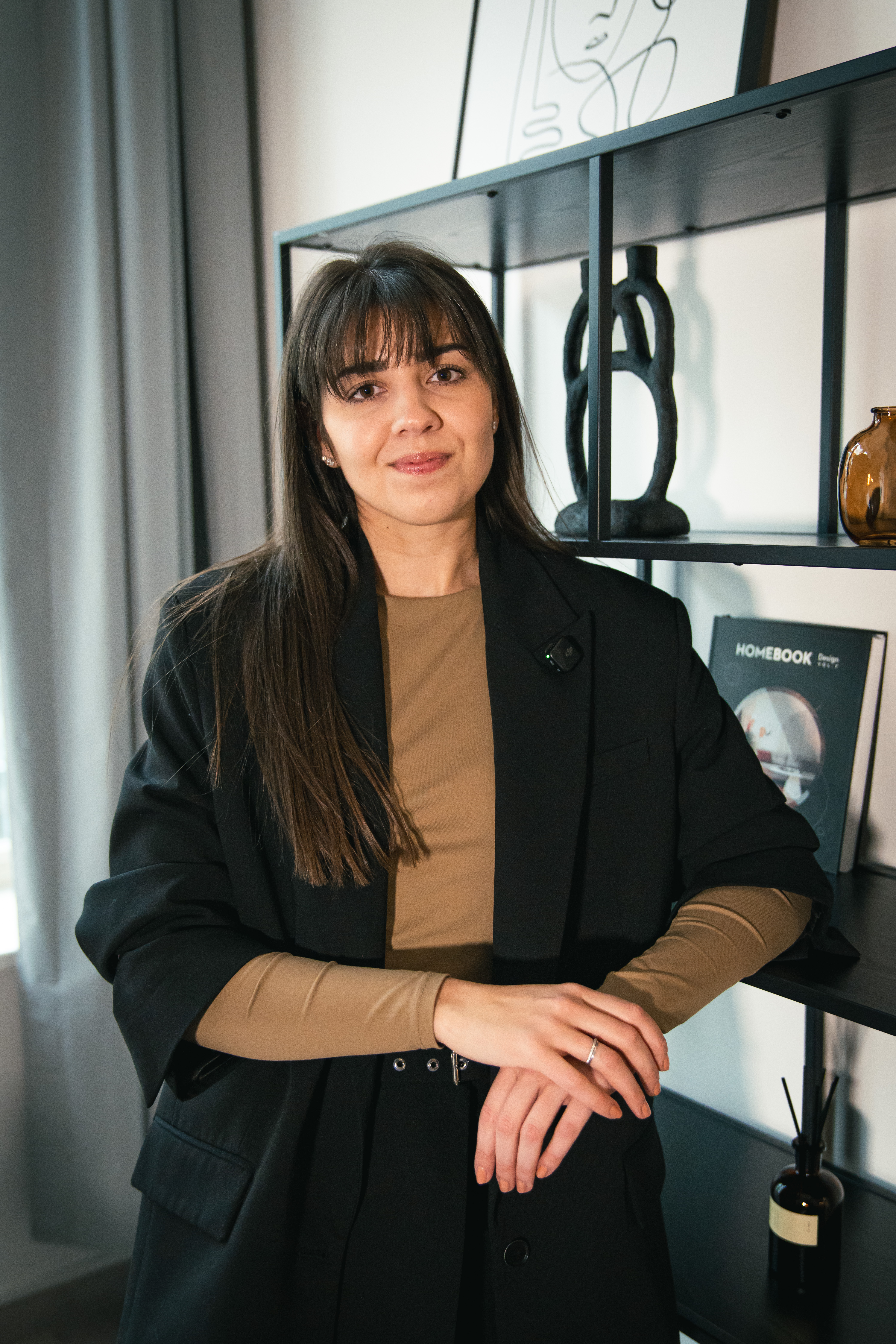 Denisa Fousová, Head of HR ve společnosti Bonami, se jemně usmívá do objektivu během natáčení série HR Vibecheck od agentury VERYGUD. Na sobě má hnědé triko s dlouhým rukávem a černé sako, na kterém má připnutý malý klopový mikrofon. Stojí v moderních kancelářích Bonami a ležérně se opírá o černý kovový regál s designovými dekoracemi a knihou.