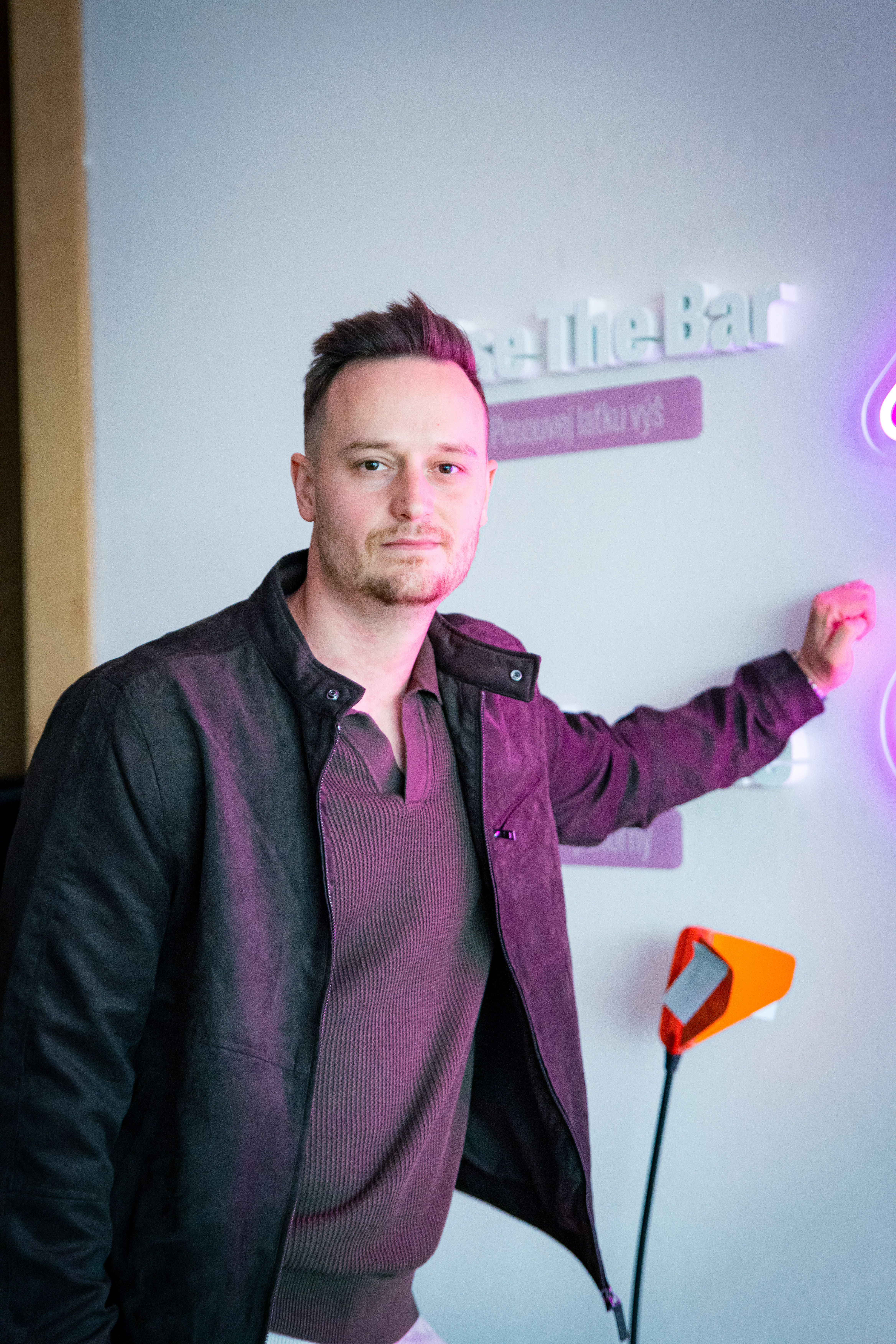 Petr Rak, Head of People ve společnosti foodora, stojí v kancelářích foodora a dívá se do objektivu. Má na sobě tmavou bundu přes texturované triko a levou rukou se ležérně opírá o bílou zeď. Na zdi za ním jsou 3D nápisy a fialově nasvícená tabulka s heslem „Posouvej laťku výš“, odrážející firemní kulturu. Foceno během natáčení rozhovoru pro sérii HR Vibecheck od agentury VERYGUD.
