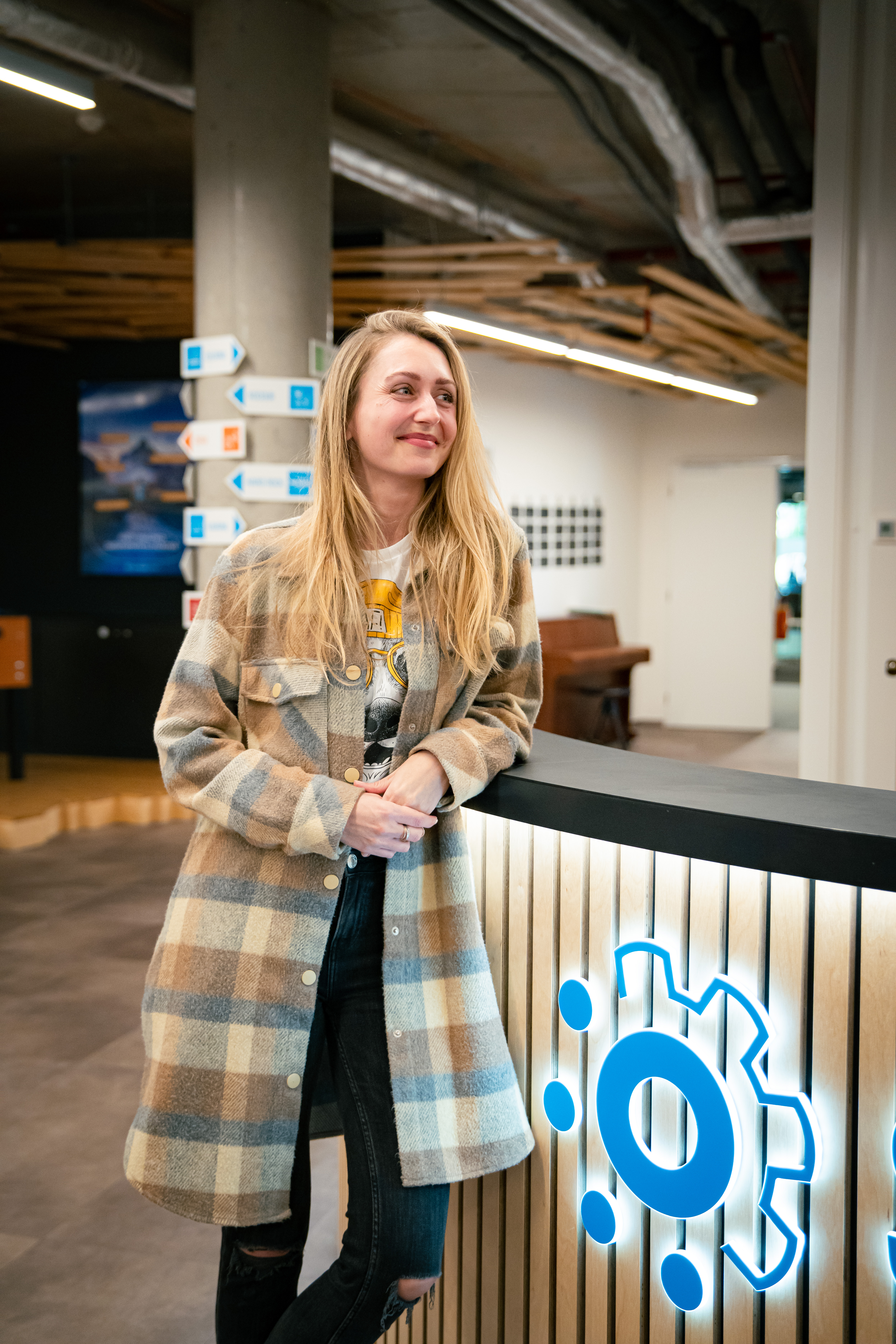 Aneta Müllerová, People Partner Lead ve společnosti Slevomat, se usmívá a ležérně se opírá o recepční pult v moderních kancelářích Slevomatu. Pult zdobí zepředu podsvícené modré logo. Aneta má na sobě dlouhou kostkovanou košilovou bundu v zemitých podzimních barvách, bílé tričko s grafickým potiskem a černé džíny s dírami na kolenou. Fotografie byla pořízena v rámci natáčení 15. dílu série HR Vibecheck produkcí VERYGUD.